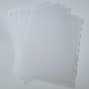 May include: A stack of translucent, rectangular plastic sheets. The sheets are arranged in a fan shape, with the edges slightly overlapping. The plastic appears to be frosted or matte, with a soft, diffused appearance. The background is a plain white surface.
