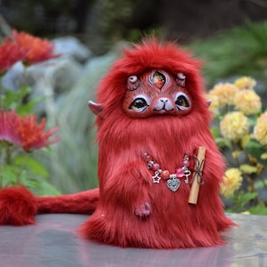 May include: A handmade red plush creature with three eyes, small horns, and a fluffy tail. It has a detailed face and is wearing a necklace with charms. The creature is holding a small wooden stick.