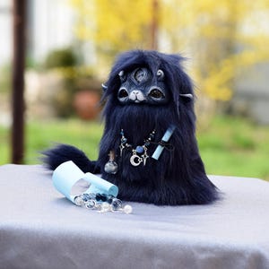 May include: A dark blue fantasy creature doll with three eyes, a furry body, and a hood. It wears a necklace with a crescent moon pendant and holds a scroll. A small bottle and beads are scattered nearby. The doll is on a grey surface.