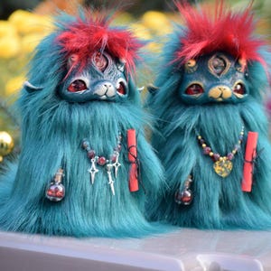 May include: Two whimsical, teal-furred creatures with red tufts of hair and three eyes. They have detailed faces, necklaces, and small potion bottles. Each holds a red scroll and a silver charm. The background is blurred with colorful foliage.