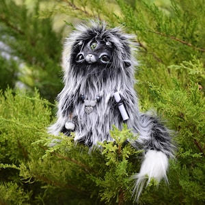 Puede incluir: Una criatura de peluche con pelo sintético largo en blanco y negro, con un tercer ojo en la frente, ojos grandes y oscuros y una nariz pequeña. Lleva un collar de cuentas con un dije de corazón y sostiene un pequeño pergamino blanco y una botella.