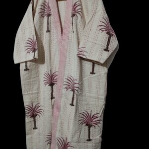 Puede incluir: Una bata de algodón blanca con estampado de palmeras rosa y marrón. La bata tiene un frente largo y abierto y un cinturón para atar.