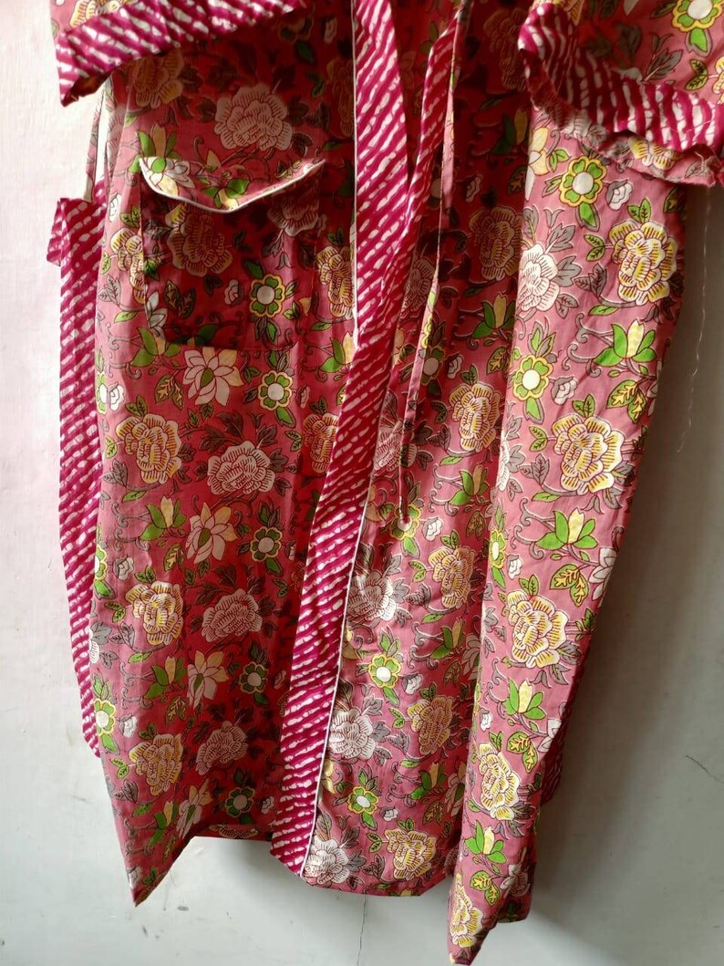 May include: A pink cotton robe with a floral print and a red and white striped tie. The robe has a white trim and two pockets.
