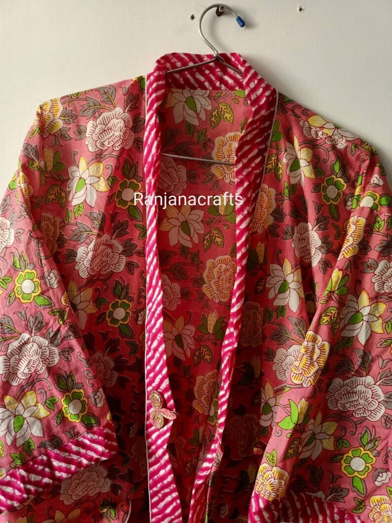May include: A pink floral print cotton robe with a pink and white striped trim. The robe has a button closure and long sleeves. The text "Ranjanacrafts" is visible on the robe.