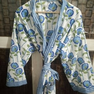 Könnte beinhalten: Eine weiße Baumwollrobe mit blauem Blumendruck und blau-weiß gestreiftem Besatz. Die Robe hat einen Bindegürtel und eine Tasche auf der linken Seite.