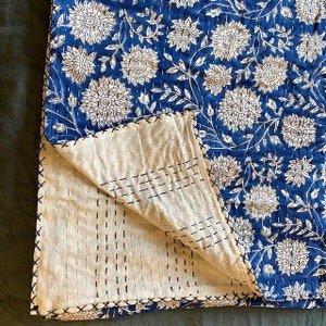 Pu&ograve; includere: Una trapunta kantha blu e bianca con un motivo floreale. La trapunta &egrave; piegata a met&agrave;, mostrando le cuciture sul retro.