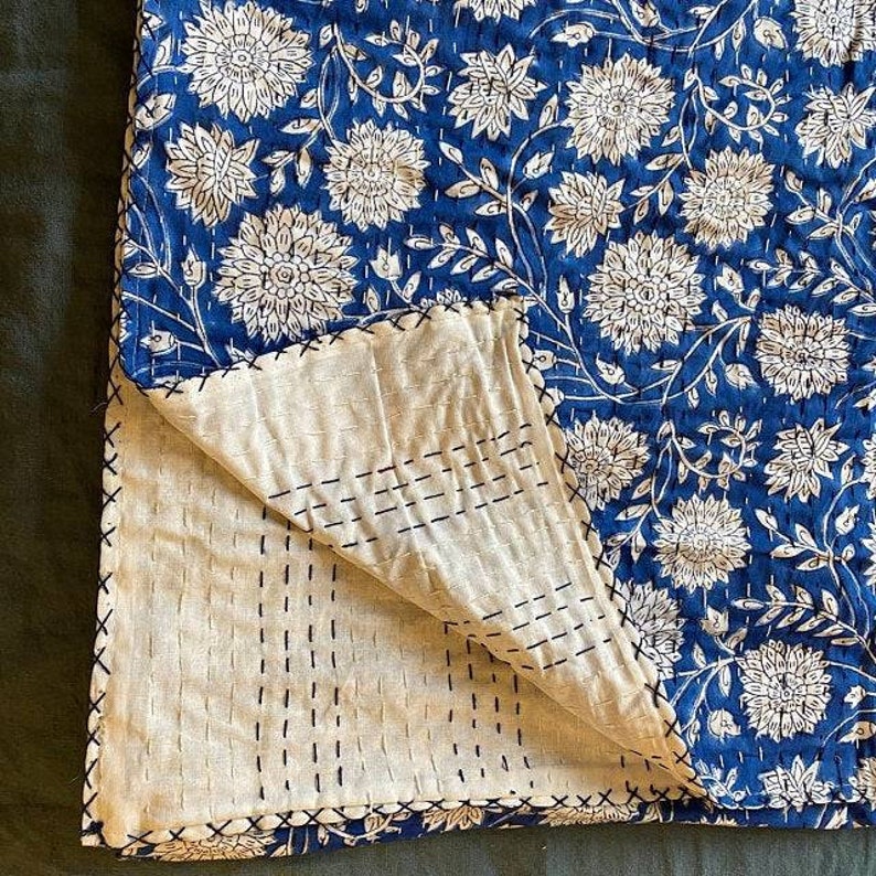 Pu&ograve; includere: Una trapunta kantha blu e bianca con un motivo floreale. La trapunta &egrave; piegata a met&agrave;, mostrando le cuciture sul retro.