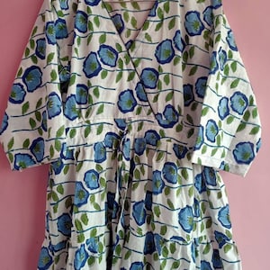 Puede incluir: Un vestido de algodón blanco con estampado floral azul y verde. El vestido tiene un escote en V y un corpiño de estilo envolvente. Cuenta con un lazo en la cintura y una falda de capas.