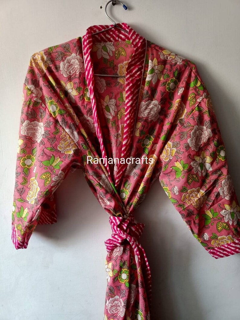 May include: A pink floral cotton robe with a white and pink striped trim. The robe has a tie belt and long sleeves. The text "Ranjanacrafts" is visible on the robe.