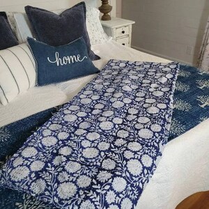 Pu&ograve; includere: Un trapunta con motivo floreale blu e bianco con sfondo bianco e fiori blu. La trapunta &egrave; piegata su un letto con cuscini blu e un cuscino con la parola "home" su di esso.