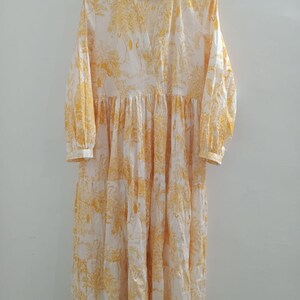 Puede incluir: Un vestido maxi blanco con estampado floral amarillo. El vestido tiene un escote en V y mangas largas.