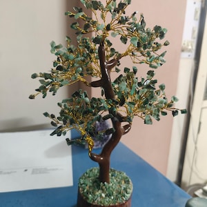May include: A decorative tree sculpture featuring a brown trunk and branches made of twisted gold-colored wire. The branches are adorned with small, tumbled green stone chips. The tree is mounted on a cylindrical base covered in green stone chips.