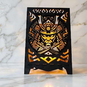 Könnte beinhalten: Eine schwarze Holzlampe mit einem lasergeschnittenen Design eines Samurai-Kriegers mit Helm. Das komplizierte Ausschnittmuster wird von innen durch ein warmes gelbes Licht beleuchtet, was einen leuchtenden Effekt auf dem dunklen Holzrahmen erzeugt.