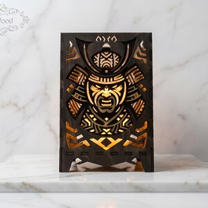 Könnte beinhalten: Eine dunkelbraune, lasergeschnittene Holzlampe mit einem komplizierten Samurai-Masken-Design, durch dessen Ausschnitte warmes Licht scheint. In der oberen linken Ecke befindet sich der Schriftzug ICSwood.
