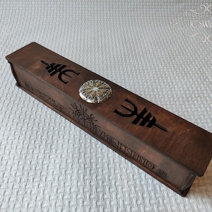 Può includere: Una scatola di legno con un design decorativo e un cerchio di metallo al centro. La scatola è incisa con rune e le parole "The First Sentinel".