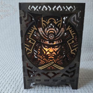 Op de afbeelding: Een houten doos met een lasergesneden ontwerp van het gezicht van een samoerai-krijger. Het ontwerp is in goud en zwart, en de doos is donkerbruin.