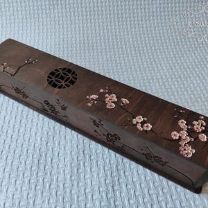 Puede incluir: Una caja rectangular de madera con acabado marrón oscuro. La caja presenta un diseño floral con flores rosas y un recorte circular. La caja mide aproximadamente 30 cm de largo.