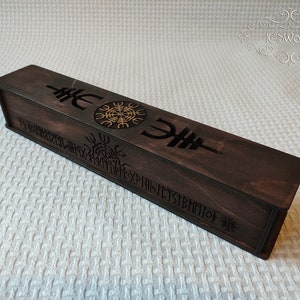 Può includere: Una scatola di legno marrone scuro con intagli intricati di rune e un simbolo. La scatola è chiusa e ha una forma rettangolare.