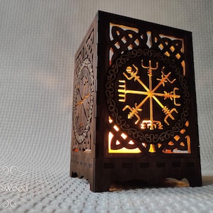 May include: A dark brown wooden lantern with intricate cut-out designs. The lantern features a circular emblem with a central starburst design and other geometric patterns. The light inside casts a warm glow through the cutouts.
