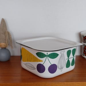 Pode incluir: Um recipiente de armazenamento quadrado de esmalte branco com tampa, apresentando uma estampa botânica colorida de uma cenoura amarela, cerejas roxas e folhas verdes. O recipiente tem uma borda preta fina e um acabamento liso.