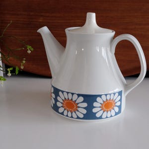 May include: White ceramic teapot with a blue band featuring a daisy pattern with orange centers. The teapot has a curved handle, a spout, and a lid with a small knob. The background is a wooden surface.