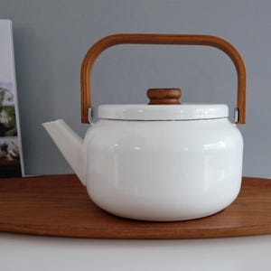 May include: A white enameled metal teapot with a rounded body, a short spout, and a flat lid featuring a small wooden knob. The teapot has a tall, arched wooden handle attached to the sides, providing a contrast to the smooth white finish.