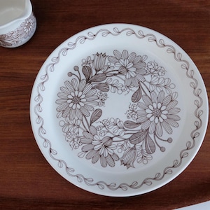 Puede incluir: Un plato de cerámica blanco con un diseño floral marrón y un borde decorativo. También se ven una crema a juego y una taza blanca pequeña. Los artículos están sobre una superficie de madera.