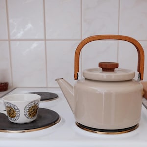 May include: A beige enamel kettle with a wooden handle and lid sits on a white stovetop. A matching teacup with a floral design rests on a burner. Kitchenware and home decor.