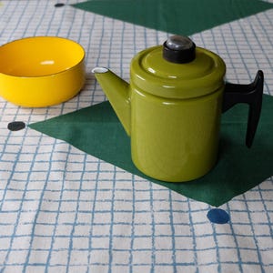 Peut inclure: Un bol jaune vif et une théière vert olive brillant avec une poignée et un bouton de couvercle noirs. La théière présente un corps cylindrique et un bec verseur pointu. Les deux objets sont posés sur un tissu blanc à carreaux bleus et losanges verts.