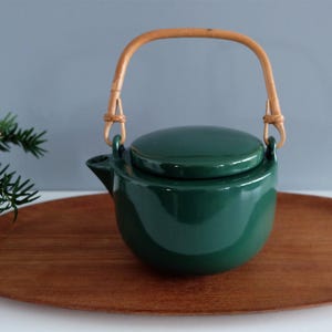 Op de afbeelding: Een donkergroene keramische theepot met een bijpassend deksel en een bamboe handvat. De theepot staat op een houten dienblad. De theepot heeft een glanzende afwerking en een afgeronde vorm. De handgreep is bevestigd met gevlochten rotan.