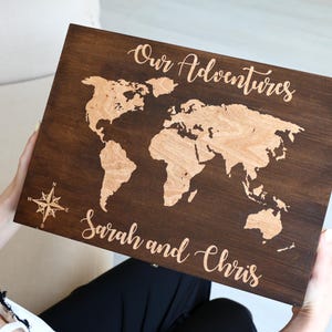 May include: A rectangular dark brown wooden board featuring a light-colored laser-engraved world map. The text "Our Adventures" is engraved at the top, and the names "Sarah and Chris" are engraved at the bottom, with a compass rose on the left.