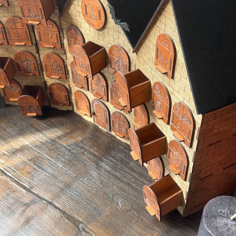 Op de afbeelding: Houten adventskalender in de vorm van een huis. Het heeft veel kleine, genummerde deuren en lades. Het huis heeft een beige buitenkant met bruine details en een zwart dak. Er staat een decoratieve vleermuis op het dak.