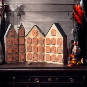 Op de afbeelding: Houten adventskalender in de vorm van drie huizen. Elk huis heeft meerdere deuren met nummers en illustraties. De huizen zijn gemaakt van lichtbruin hout met oranje deuren en zwarte daken. De kalender staat op een donkerbruin oppervlak.