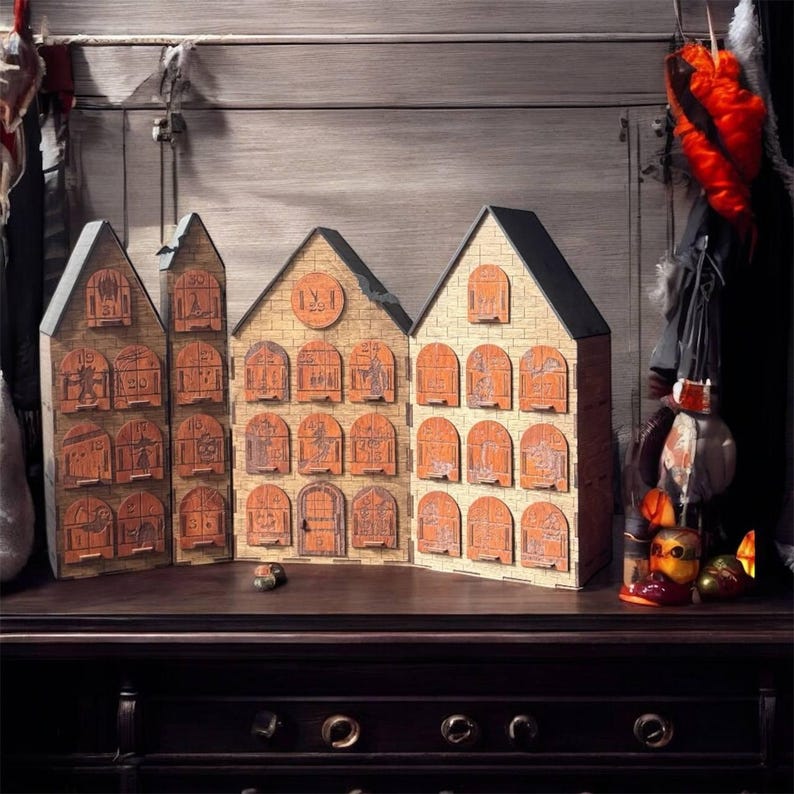Op de afbeelding: Houten adventskalender in de vorm van drie huizen. Elk huis heeft meerdere deuren met nummers en illustraties. De huizen zijn gemaakt van lichtbruin hout met oranje deuren en zwarte daken. De kalender staat op een donkerbruin oppervlak.