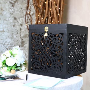 May include: A black wooden card box with intricate, openwork designs on the sides. The box has a hinged lid secured with a gold-colored lock and key. A white floral arrangement and stationery are on the table.