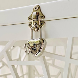 Può includere: Primo piano di un forziere bianco con un lucchetto e una chiave argentati a forma di cuore. Il lucchetto è attaccato a un fermo argentato fissato con quattro viti. Il forziere ha un motivo geometrico sul davanti.