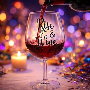 Puede incluir: Una copa de vino que se está llenando con vino tinto, con las palabras "Rise & Wine" en una fuente cursiva en la copa. Una vela encendida y luces bokeh coloridas en el fondo.