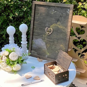Puede incluir: Una caja de madera con un diseño de corte en forma de corazón, llena de pequeños corazones de madera. La caja está abierta y se encuentra sobre una mesa blanca con un letrero que dice "Wedding Guest Book, Please Sign a Heart, Then Drop it in the Box, to Celebrate our Love and Journey Together".