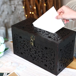 May include: Dark brown wooden card box with an intricate floral cut-out design. The box has a hinged lid with a brass-coloured clasp. A hand is inserting a white card into the slot on top. The box is on a table with a white tablecloth.