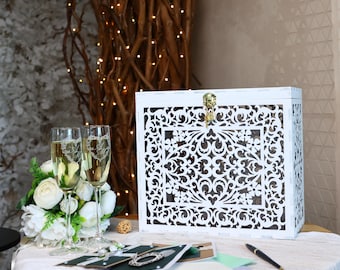 May include: A white wooden card box with intricate floral cutouts and a gold clasp. Two champagne flutes with engraved initials and a bouquet of white flowers sit nearby. A pen and cards are on the table, suggesting a wedding or celebration.