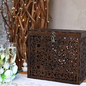 Puede incluir: Una caja de tarjetas de madera marrón oscuro con intrincados diseños florales cortados con láser. La caja tiene una tapa con bisagras y un cierre de metal con un candado. Dos copas de champán con iniciales grabadas y un ramo de flores blancas están en primer plano.