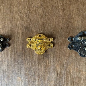 May include: Three decorative metal clasps in varying sizes and finishes. The clasps are ornate, with a dark bronze, gold, and black finish. The clasps are designed for boxes or small containers. The background is a wooden surface.