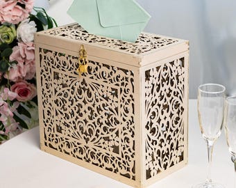 May include: A cream-colored wooden card box with intricate cut-out designs. A hand is placing a mint green envelope into the box. The box has a gold-colored lock. Champagne flutes and stationery are on the table.