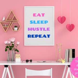 Pode incluir: Um cartaz motivacional emoldurado em branco com o texto "Eat Sleep Hustle Repeat" em letras rosa, azul e azul claro. O cartaz está pendurado em uma parede rosa com uma mesa branca na frente. Há dois corações de papel rosa pendurados no teto acima do cartaz.
