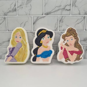 May include: Three bath bombs shaped like Disney princesses. The bath bombs are white with colourful images of Jasmine, Belle, and Rapunzel.