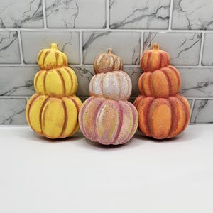 May include: Three stacked pumpkin-shaped bath bombs arranged in a row. The left bath bomb is yellow with orange accents, the centre one is a pale pink and white blend, and the right one is orange with darker orange shading. Each has a ribbed texture.
