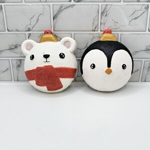 May include: Two bath bombs shaped like a polar bear and a penguin. The polar bear is wearing a red scarf and a gold hat. The penguin is wearing a black hat. Both bath bombs are white with black eyes and a black nose.