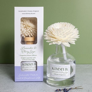 May include: A white wooden flower diffuser with a glass bottle filled with clear liquid. The bottle has a label that reads "KIMMYB. Lavender & Clary Sage Diffuser Fragrance". The diffuser is packaged in a white box with a lavender and white design. The box reads "Handmade in Sussex, England www.Kimmy-B.com Lavender & Clary Sage Fragrance Flower Diffuser English Lavender with a hint of Clary Sage KIMMYB. Lavender & Clary Sage Diffuser Fragrance Sustainable Products made to Complement Life"