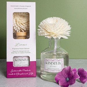 May include: A clear glass fragrance diffuser with a white flower and a box. The box is white with a pink stripe and the text "L'amour Fragrance Flower Diffuser" and "Sustainable Products made to Complement Life". The diffuser has a white flower and a purple flower.