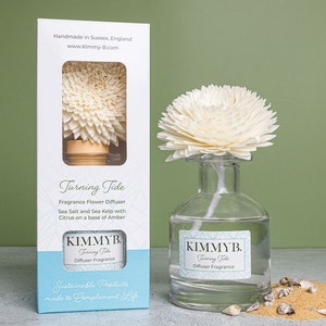 May include: A white ceramic flower diffuser with a glass bottle filled with clear liquid. The diffuser is labelled "KIMMYB. Turning Tide Diffuser Fragrance" and the box it comes in says "Sustainable Products made to Complement Life".